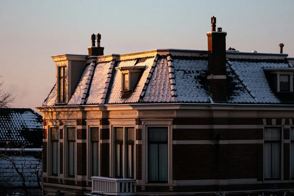 Een herenhuis met een besneeuwd dak, dakkapellen en schoorstenen in het eerste zonlicht van een winterochtend