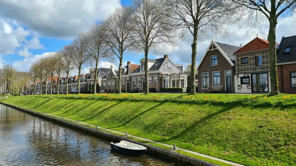 Een rij Nederlandse huizen achter een grasdijk langs een vaart, met kale bomen en een roeiboot aan de kant